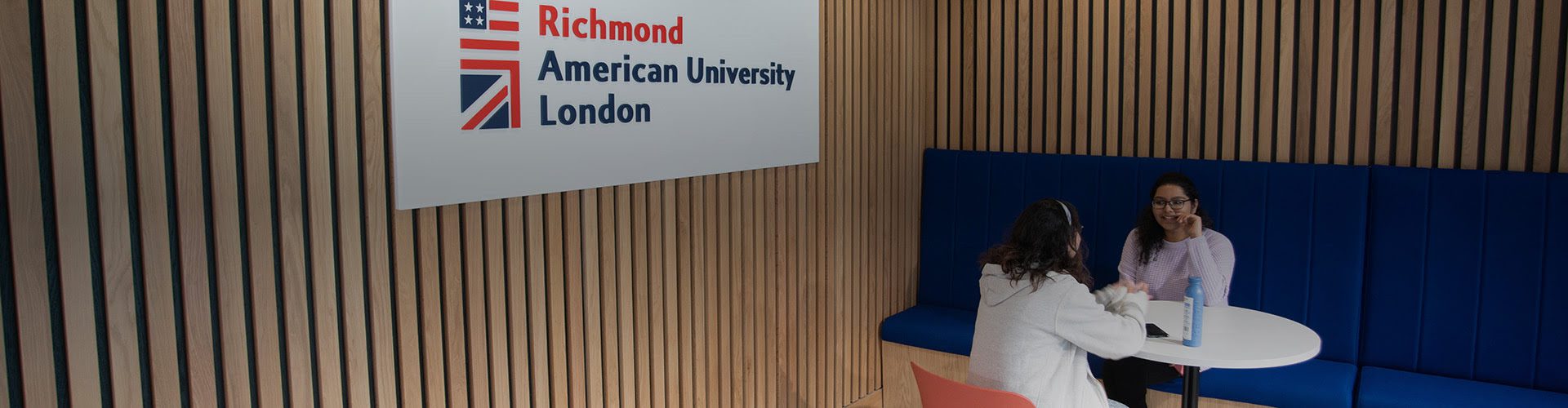 Two people are seated opposite each other at a white table inside a modern room with wooden slats and a sign for 玩偶姐姐 American University London.